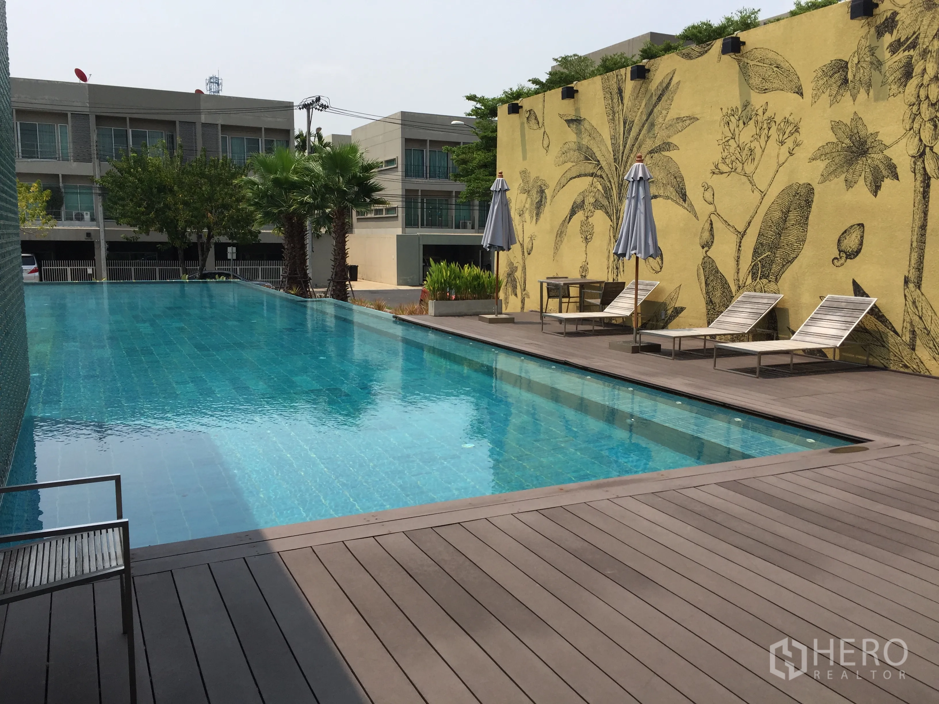 Townhouse for sale in Suan Luang, Bangkok - Outdoor swimming pool with sun loungers, palm trees, and a decorative mural wall.