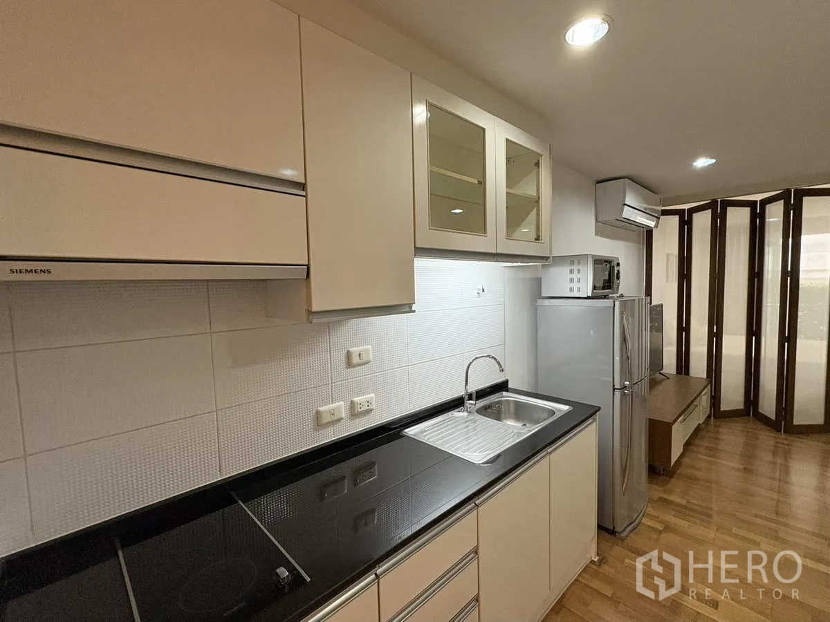 Condominium for rent in Khlong Toei, Bangkok - Fitted kitchen with glossy cabinets, electric hob, sink and black countertop.