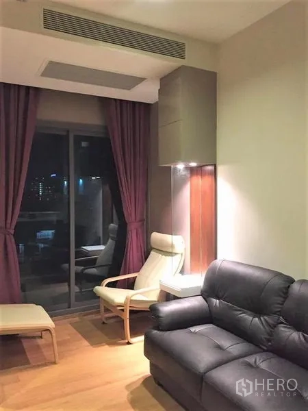 Condominium for rent or sale in Watthana, Bangkok - Living area with leather sofa, lounge chair and sliding glass door to the balcony at night.