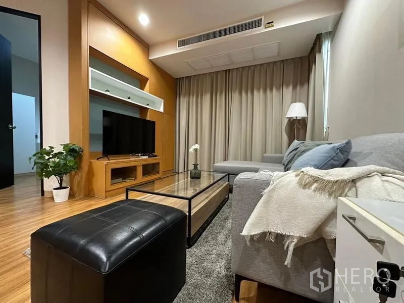 Condominium for rent in Khlong Toei, Bangkok - Warm living room with L-shaped gray sofa, glass coffee table, wood media wall and floor-to-ceiling curtains.