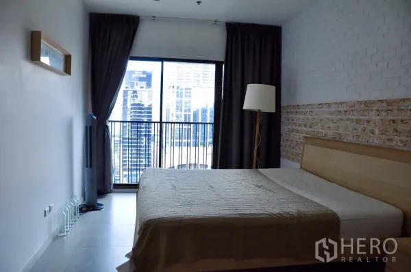 Condominium for rent in Bangkok - Bedroom with large window and balcony railing showing city skyline, dark curtains and a brick feature wall.