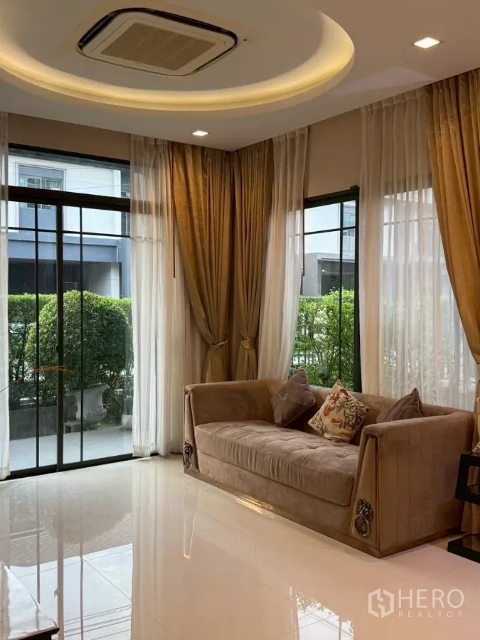 Detached House for rent or sale in Bang Phli, Samut Prakan - Bright living room with floor‑to‑ceiling sliding glass doors, sheer curtains, a velvet sofa and a ceiling cassette air conditioner.