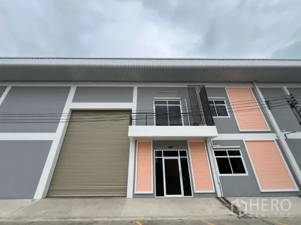 Factory for rent in Phra Samut Chedi, Samut Prakan - Street-facing factory exterior with tall shutter door and glass entry to the two-story office.