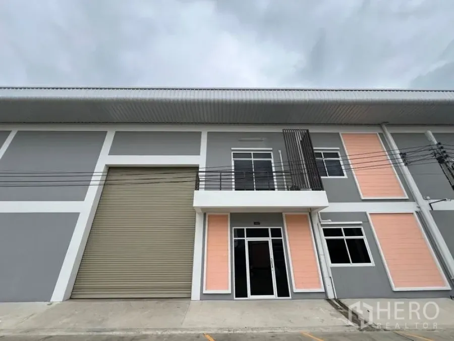Factory for rent in Phra Samut Chedi, Samut Prakan - Street-facing factory exterior with tall shutter door and glass entry to the two-story office.