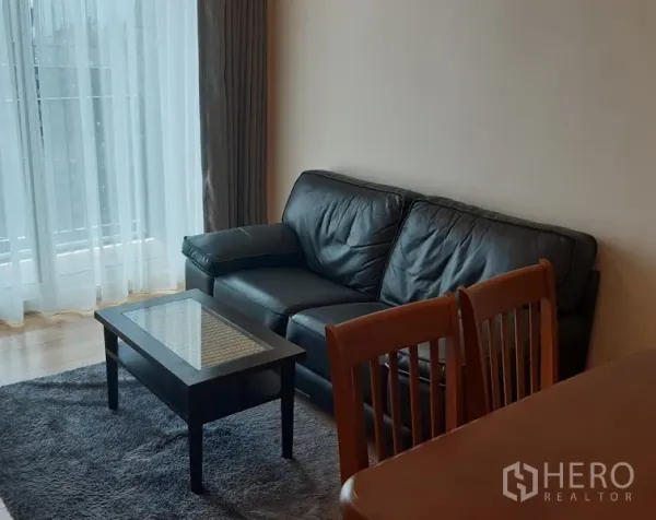 Condominium for rent in Khlong Toei, Bangkok - Cozy living area with black leather sofa, glass-top coffee table, dining chairs, and a floor-to-ceiling window with sheer curtains.