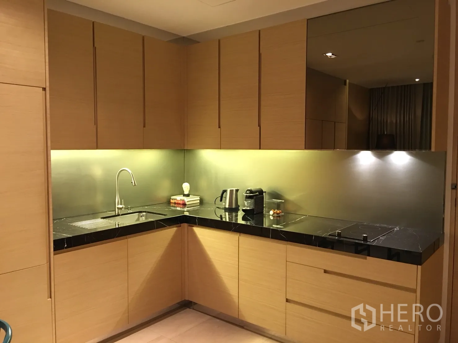 Condominium for rent or sale in Bang Rak, Bangkok - L-shaped kitchen with under-cabinet lighting, stainless backsplash, sink and electric cooktop.