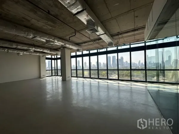 办公空间 出租在Watthana, Bangkok - 开放式办公室，落地窗面向公园与曼谷天际线，裸露天花与玻璃隔断可见。