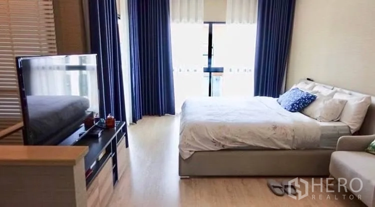 Townhouse for rent or sale in Suan Luang, Bangkok - Bedroom view showing the bed opposite a TV stand and a balcony with blue drapes.