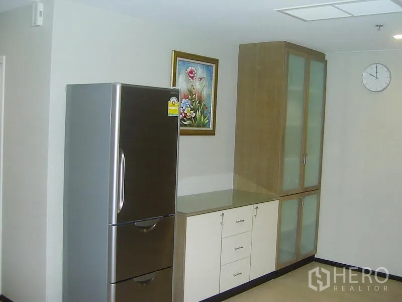 Condominium for rent in Khlong Toei, Bangkok - Kitchen corner showing stainless refrigerator and built-in cabinets with frosted glass doors and wall art.
