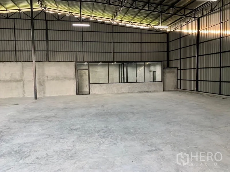 Warehouse for rent in Khan Na Yao, Bangkok - Spacious warehouse interior with high steel roof, concrete floor, and a glass-partitioned office along the back wall.