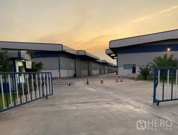 Warehouse for rent in Lat Krabang, Bangkok - Secure gated entrance to a warehouse compound with sliding gate and wide driveway at sunset.