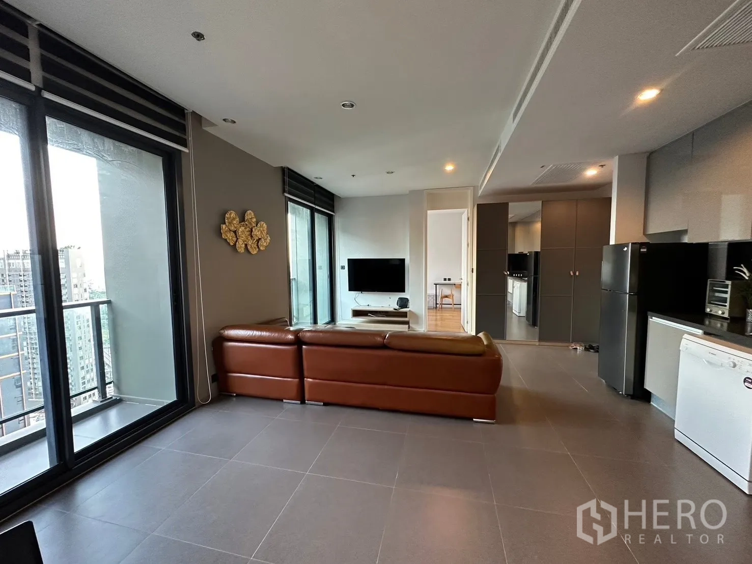 Condominium for rent or sale in Chatuchak, Bangkok - Open-plan living room with brown leather sectional, TV, and balcony access.