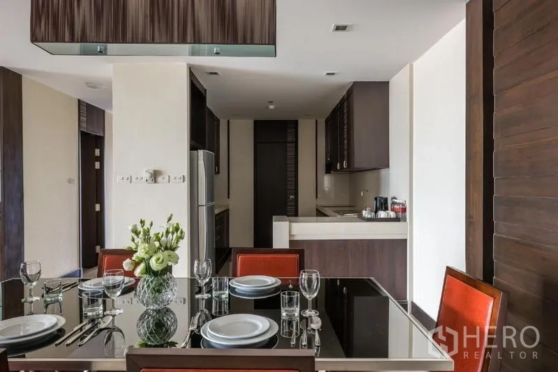Apartment for rent in Khlong Toei, Bangkok - Modern dining area with glass-top table set and view to a sleek kitchen with dark wood cabinets.
