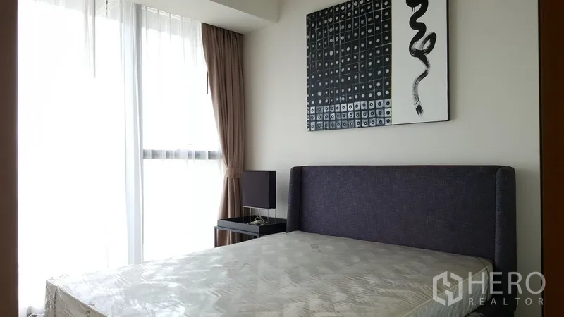 Condominium for rent in Sathon, Bangkok - Bright secondary bedroom with upholstered bed, bedside table, artwork, and sheer curtains.