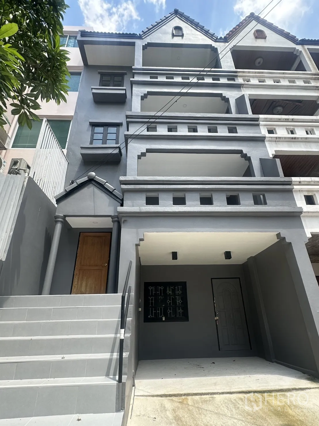 ทาวน์เฮาส์ for rent in Watthana, Bangkok - ด้านหน้าอาคารทาวน์เฮ้าส์ 4 ชั้นสีเทา มีระเบียงหลายชั้น บันไดทางเข้า และที่จอดรถในร่ม