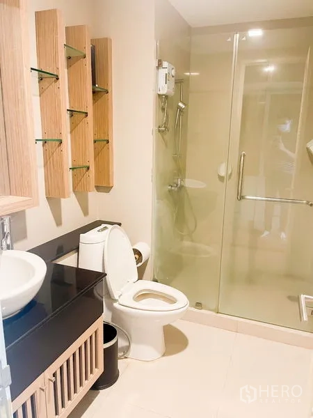Condominium for rent in Khlong Luang, Pathum Thani - Contemporary bathroom with glass-enclosed shower, vanity with basin, and wall shelving.