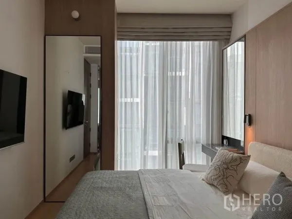 Condominium for rent in Pathum Wan, Bangkok - Bedroom with floor-to-ceiling sheer curtains, wall-mounted TV, vanity desk and mirror filled with natural light.