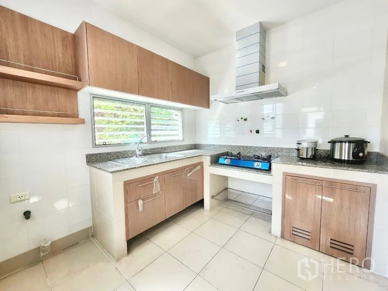 独立式住宅 出租在Prawet, Bangkok - 明亮的L形厨房，木质橱柜、燃气灶、抽油烟机与侧窗通风。