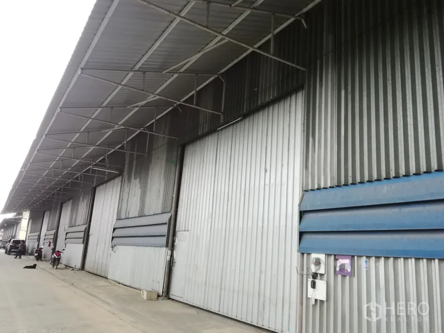 Warehouse for rent in Lam Luk Ka, Pathum Thani - Corrugated metal warehouse facade with a long covered loading bay and multiple large sliding doors in an industrial row.