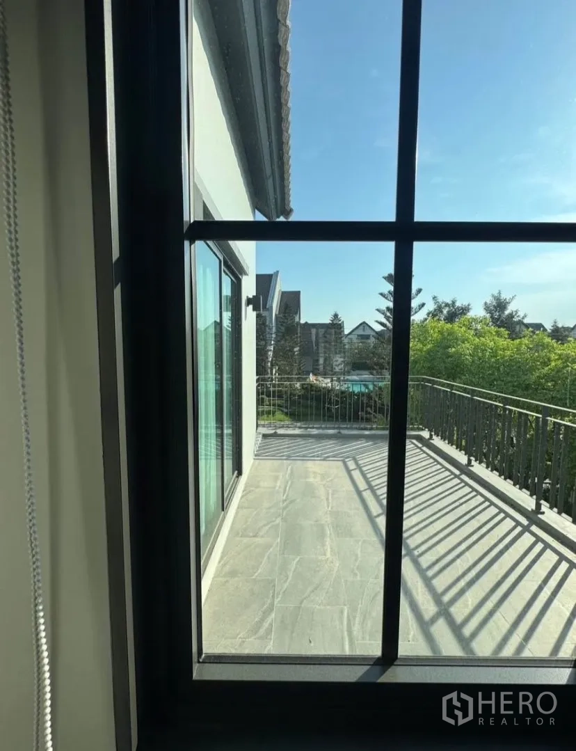 Detached House for rent or sale in Lat Krabang, Bangkok - View through a window to an upper-floor balcony with metal railing and neighborhood greenery under a blue sky.