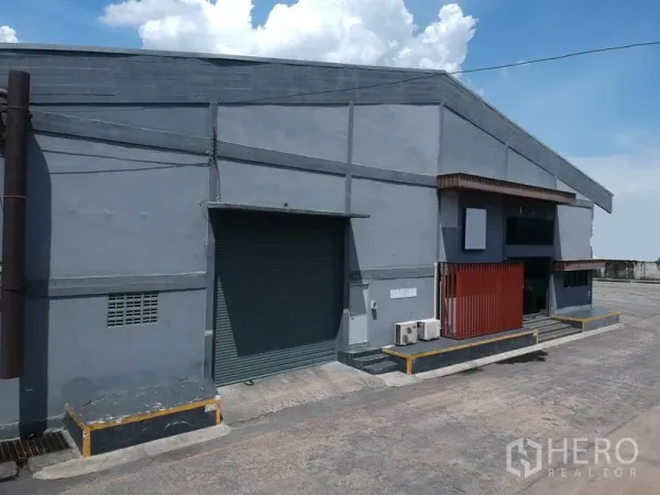Factory for rent in Phra Pradaeng, Samut Prakan - Grey factory facade featuring a loading dock, ramp, and roller shutter door under a clear sky.