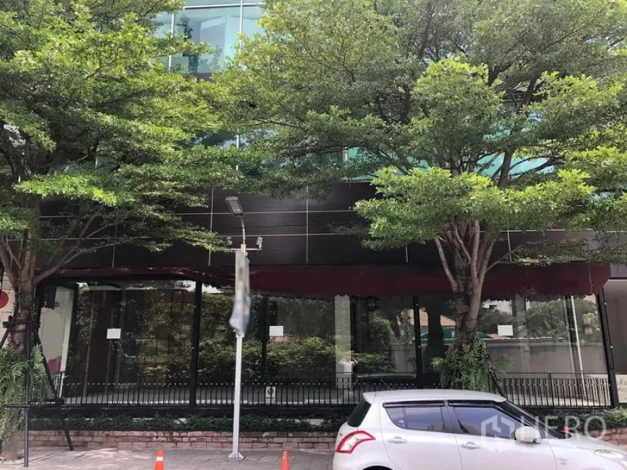 Retail Space for rent in Watthana, Bangkok - Street-facing glass facade with red awning, mature trees, and parking in front.