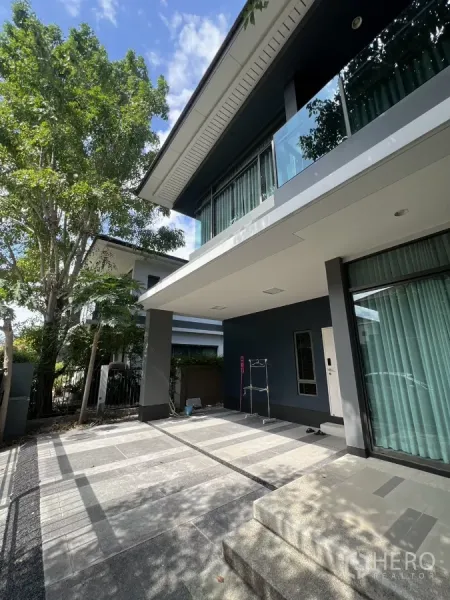 Detached House for rent in Bang Kapi, Bangkok - Covered carport and front patio beneath a cantilevered balcony with greenery around the home.