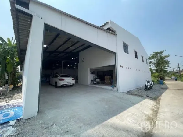 Warehouse for sale in Lam Luk Ka, Pathum Thani - White warehouse exterior with large roll‑up door, driveway and covered loading bay for vehicles.