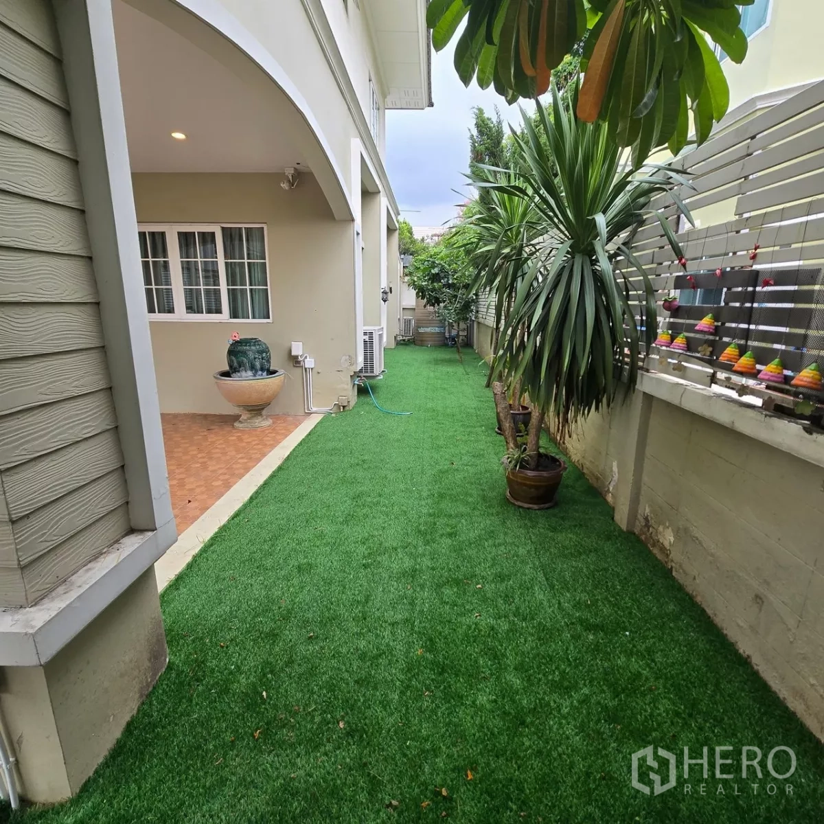 Detached House for rent in Mueang Pathum Thani, Pathum Thani - Side yard with artificial turf and potted tropical plants beside the home.