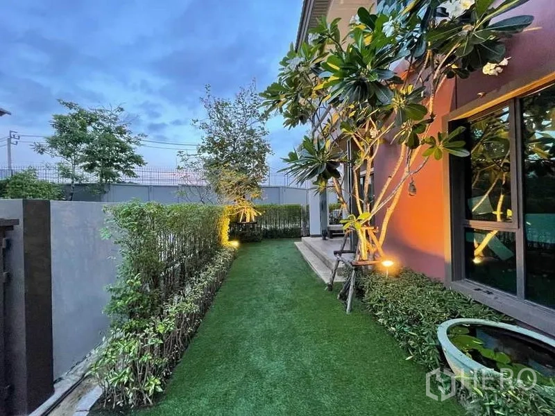 บ้าน ขายในPak Kret, Nonthaburi - สวนข้างบ้านปูหญ้าเทียม แนวพุ่มไม้และต้นลีลาวดีในช่วงพลบค่ำ