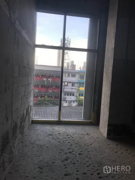 Retail Space for rent in Khlong Toei, Bangkok - Narrow space with a full-height window overlooking city buildings and a raw concrete floor.