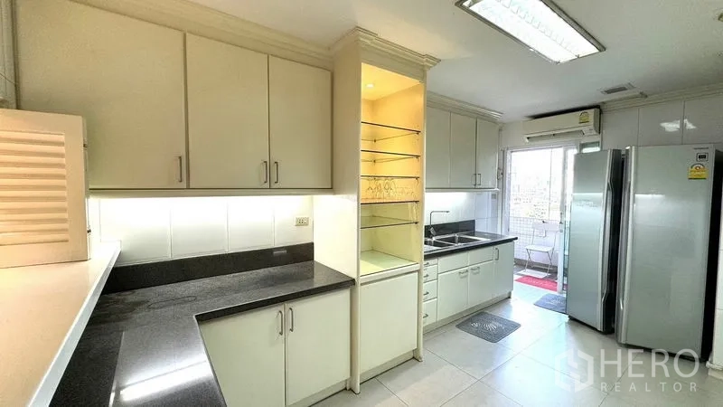 Condominium for rent or sale in Watthana, Bangkok - Bright U-shaped kitchen with cream cabinets, black countertops, double sink, balcony door, and double-door fridge.