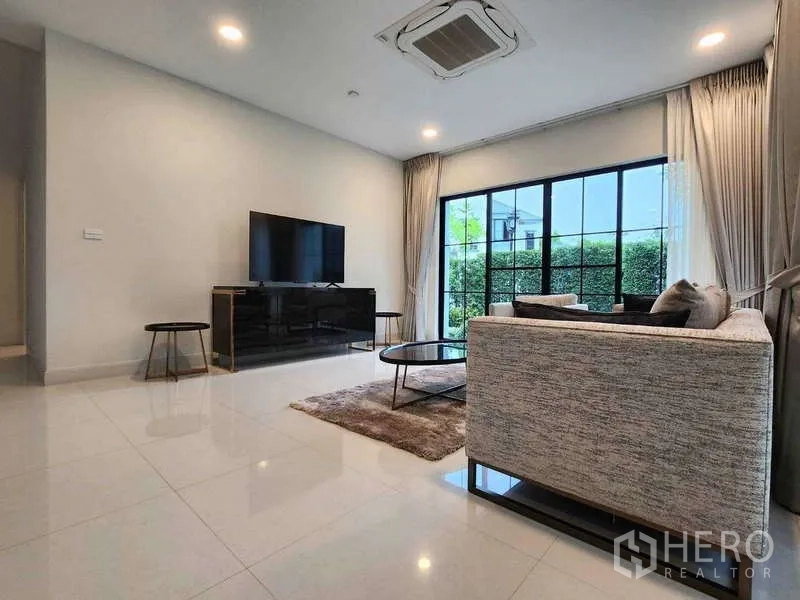 Detached House for rent in Saphan Sung, Bangkok - Lounge seating facing TV with garden views through large glass doors and full-height curtains.