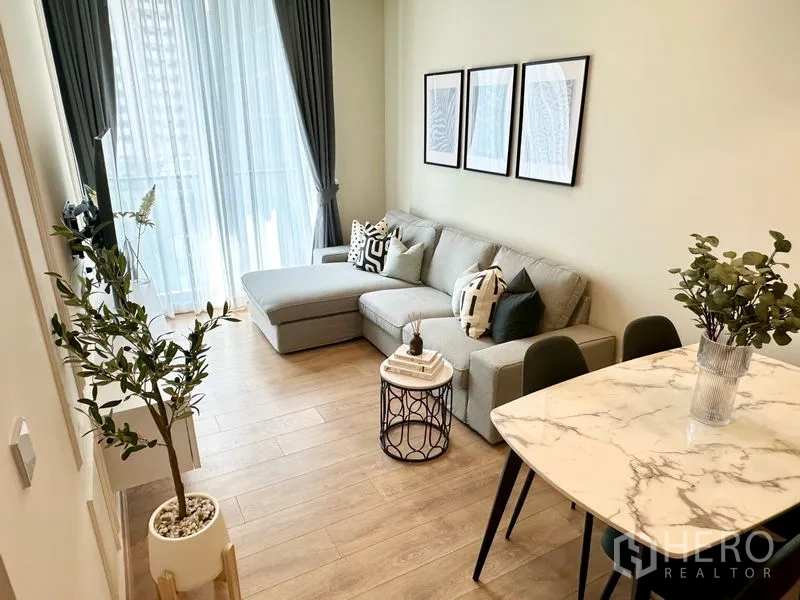 公寓 出租在Watthana, Bangkok - 明亮的公寓客厅，灰色转角沙发、石纹餐桌与落地窗帘，位于素坤逸。