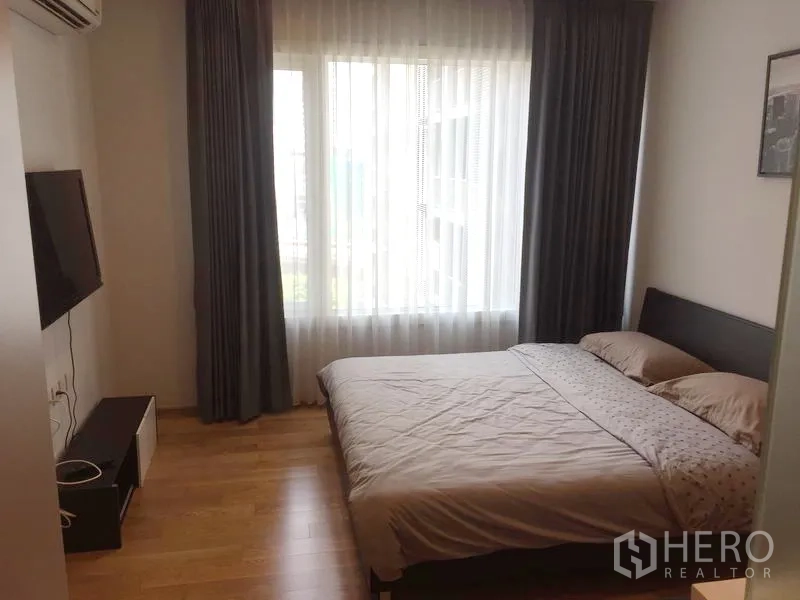 Condominium for rent in Khlong Toei, Bangkok - Bright bedroom with double bed, wall‑mounted TV and large window with sheer curtains.