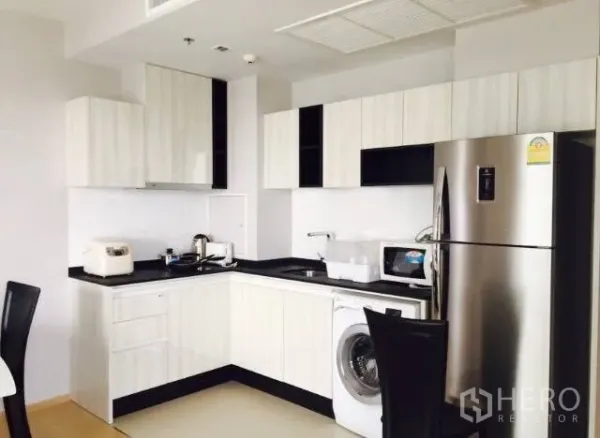 Condominium for rent in Watthana, Bangkok - Modern L-shaped kitchen with white cabinets, black countertop, washing machine, microwave and stainless refrigerator.
