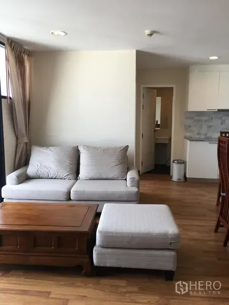 Condominium for rent in Watthana, Bangkok - Bright living room with grey sofa, ottoman and wooden coffee table beside an open kitchen in an Ekkamai condo.