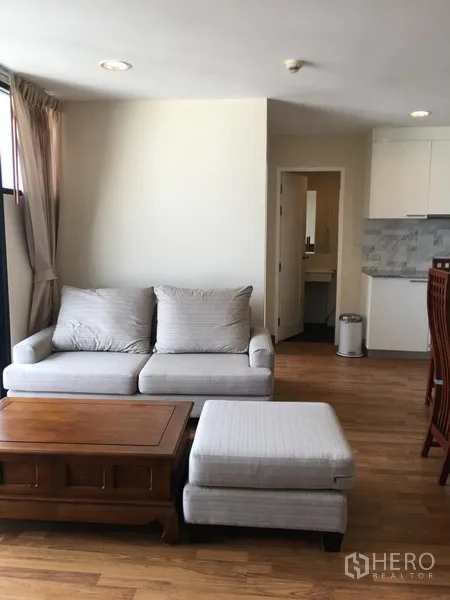 Condominium for rent in Watthana, Bangkok - Bright living room with grey sofa, ottoman and wooden coffee table beside an open kitchen in an Ekkamai condo.