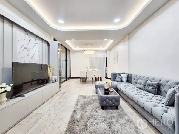 Townhouse for rent in Huai Khwang, Bangkok - Bright open-plan living room with tufted gray sofa, marble-look floors and TV wall.