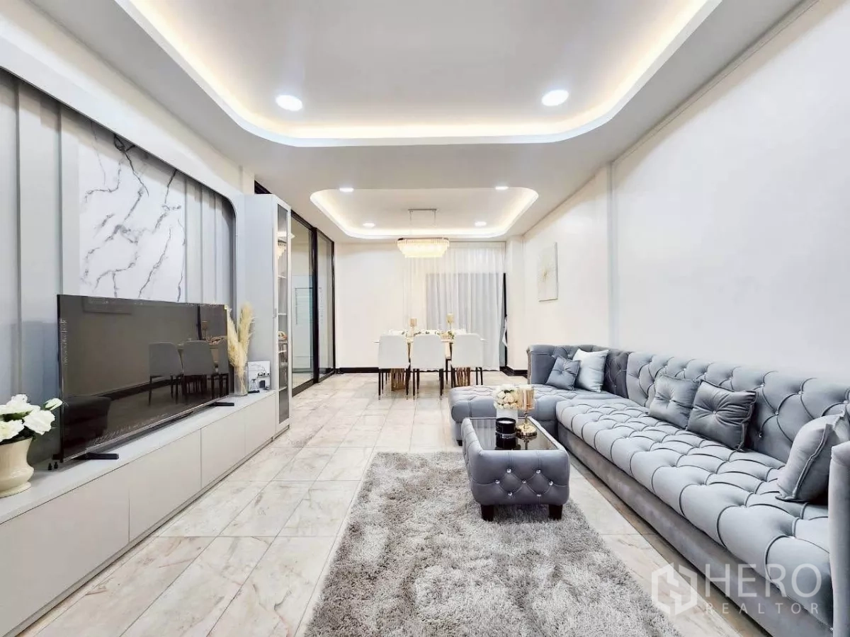 Townhouse for rent in Huai Khwang, Bangkok - Bright open-plan living room with tufted gray sofa, marble-look floors and TV wall.