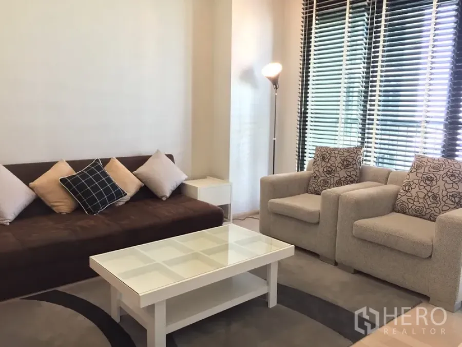 Condominium for rent in Watthana, Bangkok - Cozy living room with sofa, two armchairs, glass-top coffee table, floor lamp and large windows with blinds.