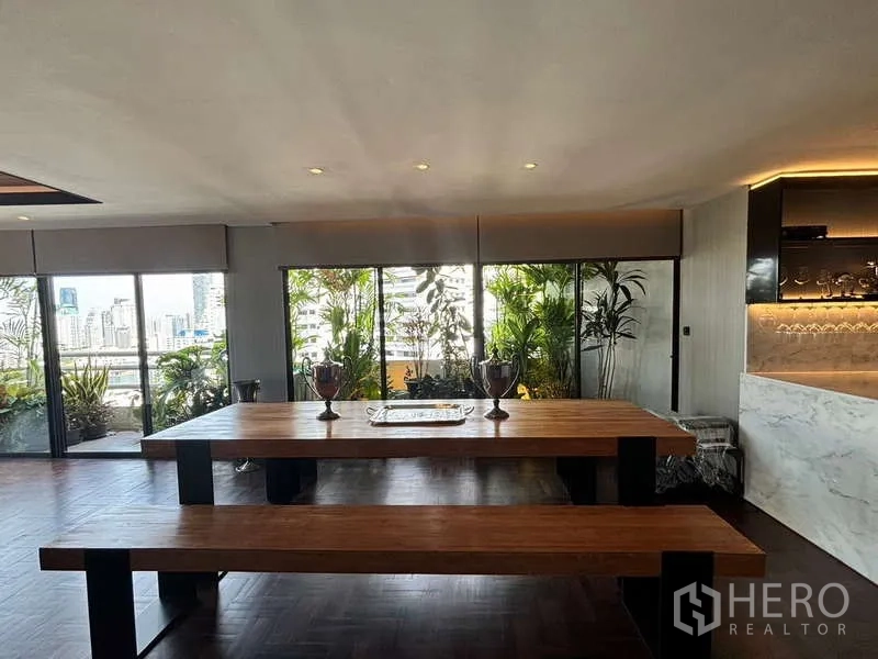 Condominium for rent in Watthana, Bangkok - Dining area with long wooden table beside floor-to-ceiling windows and balcony plants.