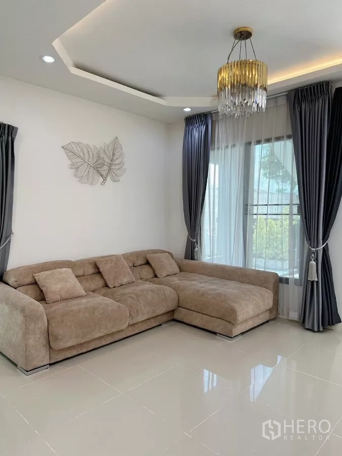 联排别墅 出租在Bang Phli, Samut Prakan - 明亮客厅，L形沙发，落地窗与纱帘，配水晶吊灯。