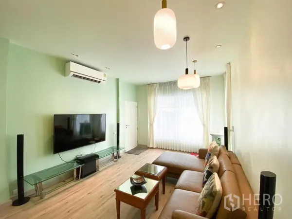 Detached House for rent in Bang Kapi, Bangkok - Bright living room with mint walls, L-shaped leather sofa, wall-mounted TV, pendant lights, and sheer curtains.
