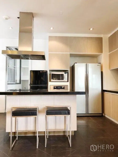 Condominium for rent in Khlong Toei, Bangkok - Kitchen island with two bar stools, built-in oven and microwave, and side-by-side refrigerator.