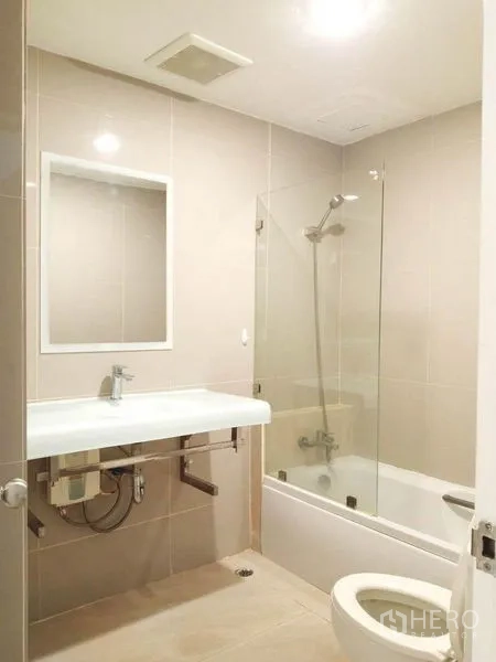 Condominium for rent in Khlong Toei, Bangkok - Clean bathroom with bathtub, glass shower screen, wall mirror and vanity in the condo.