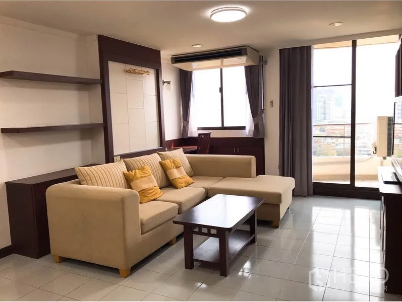 Condominium for rent or sale in Watthana, Bangkok - Living area with beige sectional, coffee table and floor-to-ceiling glass opening to city-view balcony.