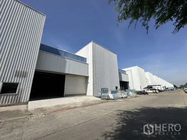 Factory for rent in Khlong Luang, Pathum Thani - Row of modern white factory warehouses with wide driveway and loading bays in Khlong Luang under a clear blue sky.