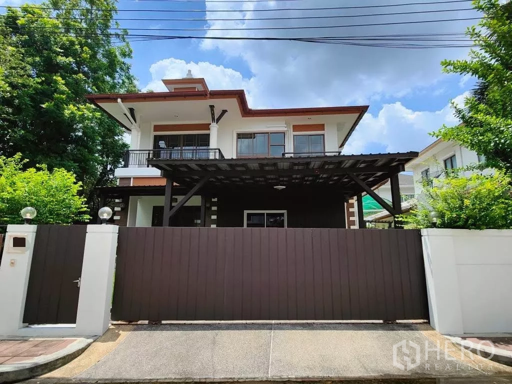 独立式住宅 出租或出售在Mueang Nonthaburi, Nonthaburi - 两层现代独栋别墅，配有大型实木大门与有顶车棚，蓝天白云下。
