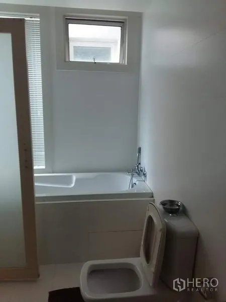 Condominium for rent in Ratchathewi, Bangkok - White bathroom featuring a bathtub beneath a small window and a frosted sliding door.